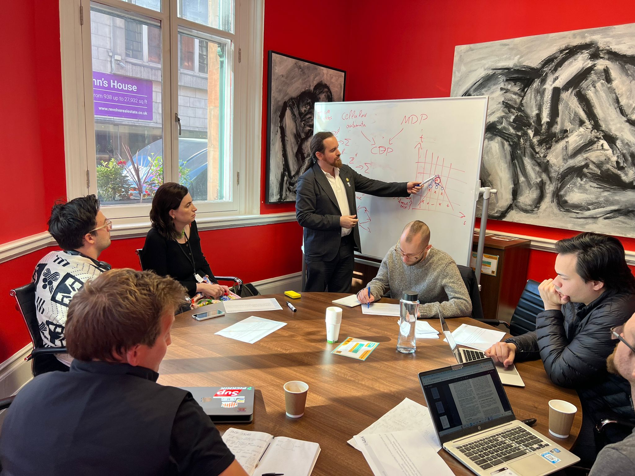A photo of a group of people sitting around the table and looking at davidad writing on a whiteboard.
